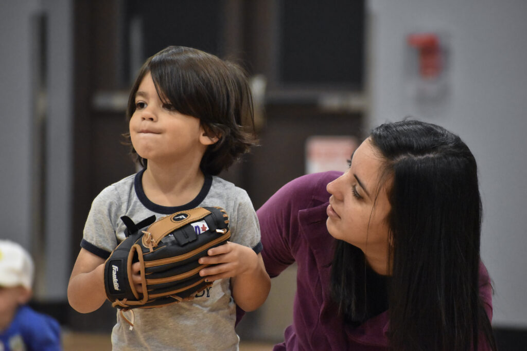 A mother kneeled down behind her son, looking at him while her son is looking to the left holding a baseball glove.