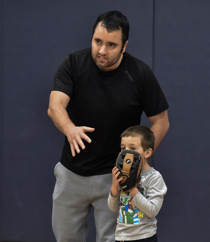 A father looking to the left while motioning with his hand, and his son standing next to him with his baseball glove covering his face, only showing his eyes.