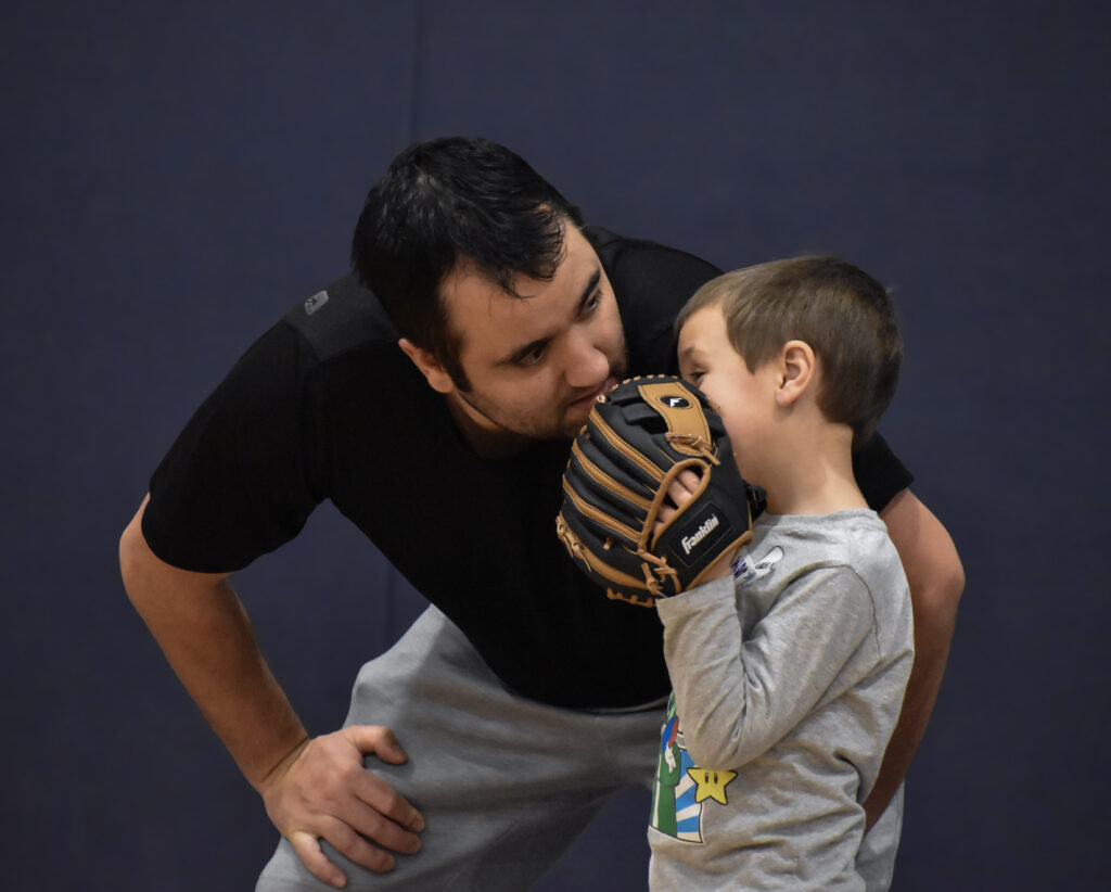 A father leaned down listening to his son while his son looks over at his father while holding his baseball glove in front of his mouth.