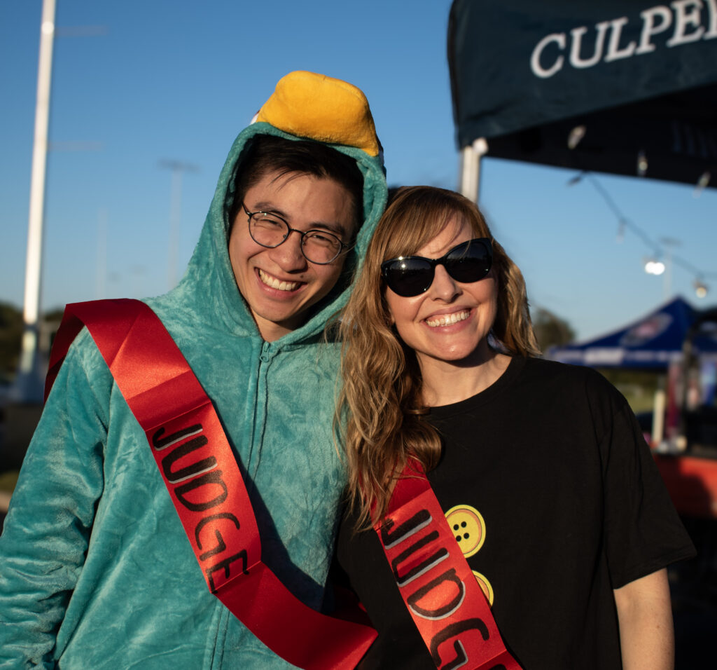 A man and a woman in costumes posing and smiling for the camera while wearing sashes that read, "Judge".