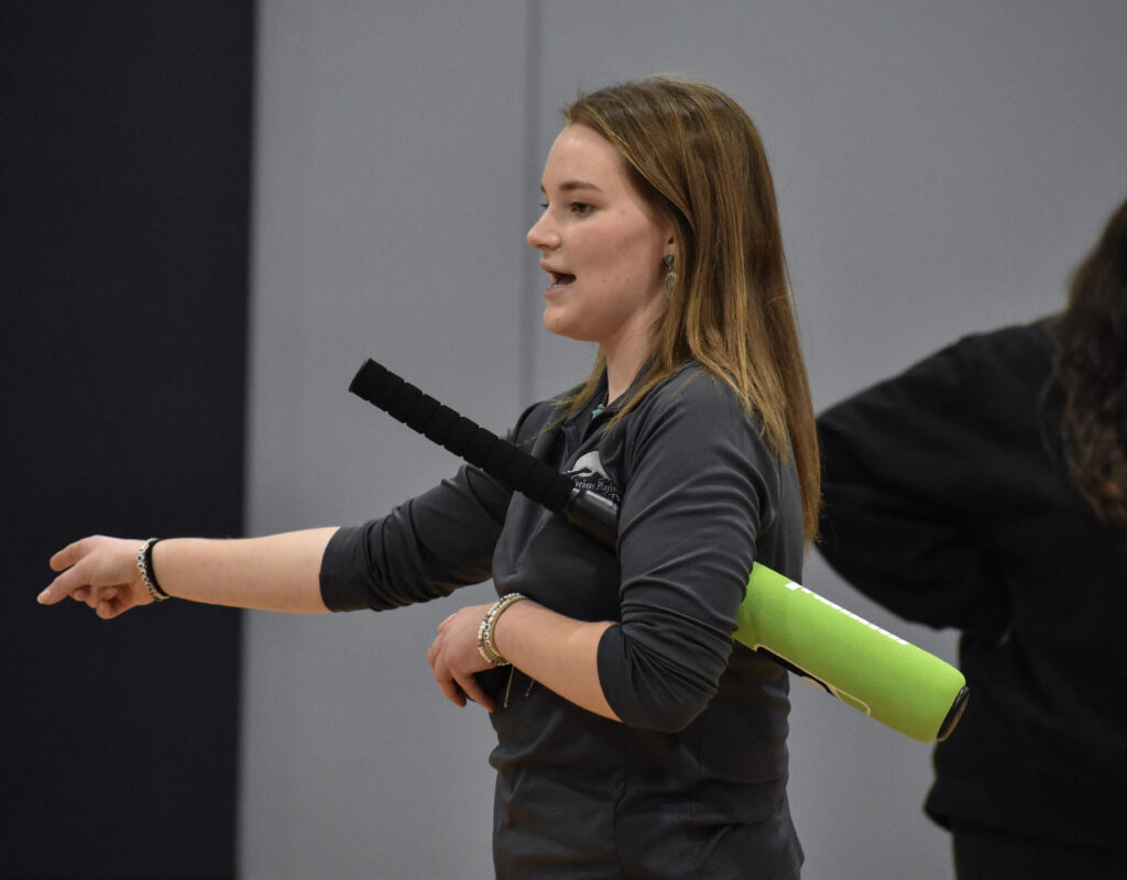 A woman instructing and holding a baseball bat under her arm while pointing in front of her.