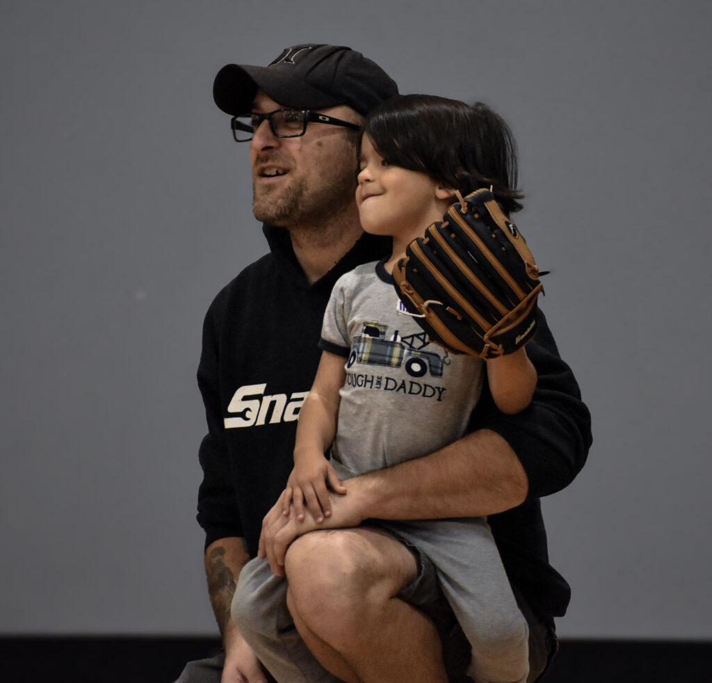 A father kneeled down with his son sitting on his propped leg, while the son his holding a baseball glove.