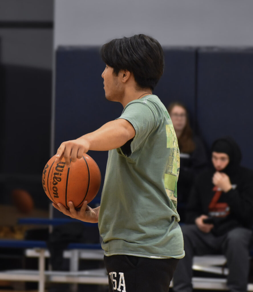 Man standing and facing to the left while holding a basketball and pointing towards the camera.